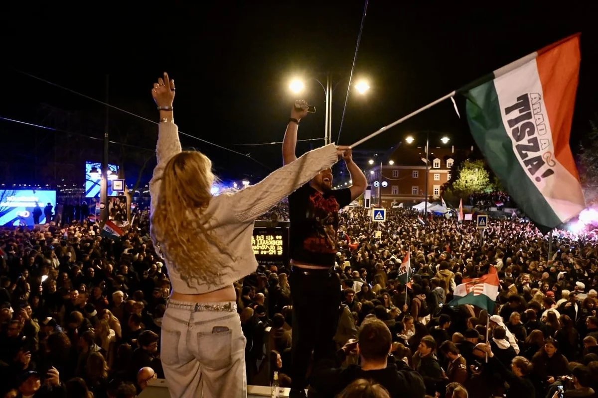People celebrating at night amid a large crowd, with a Hungarian Tisza flag raised above them